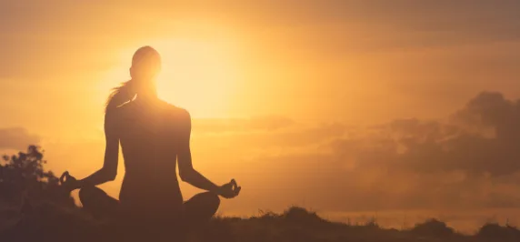 Silhueta de uma pessoa sentada em posição de meditação, em contraluz com o pôr do sol.