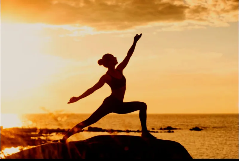 Silhueta de uma pessoa numa posição de guerreiro de Yoga, sobre uma rocha junto ao mar, ao pôr do sol.